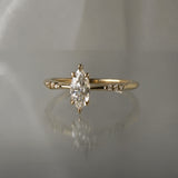 A delicate hand-carved one of a kind "Ilona" style engagement ring by Laurie Fleming Jewellery. The Ring features a 0.5ct marquise cut diamond, a tapered knife-edge band, and small diamond accents on the band shoulders. The ring is situated against a medium grey background.