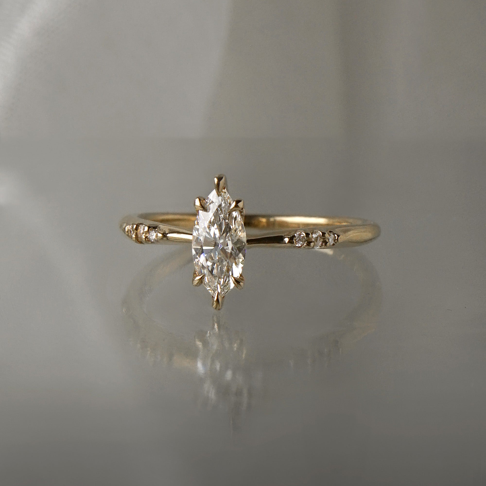 A delicate hand-carved one of a kind "Ilona" style engagement ring by Laurie Fleming Jewellery. The Ring features a 0.5ct marquise cut diamond, a tapered knife-edge band, and small diamond accents on the band shoulders. The ring is situated against a medium grey background.