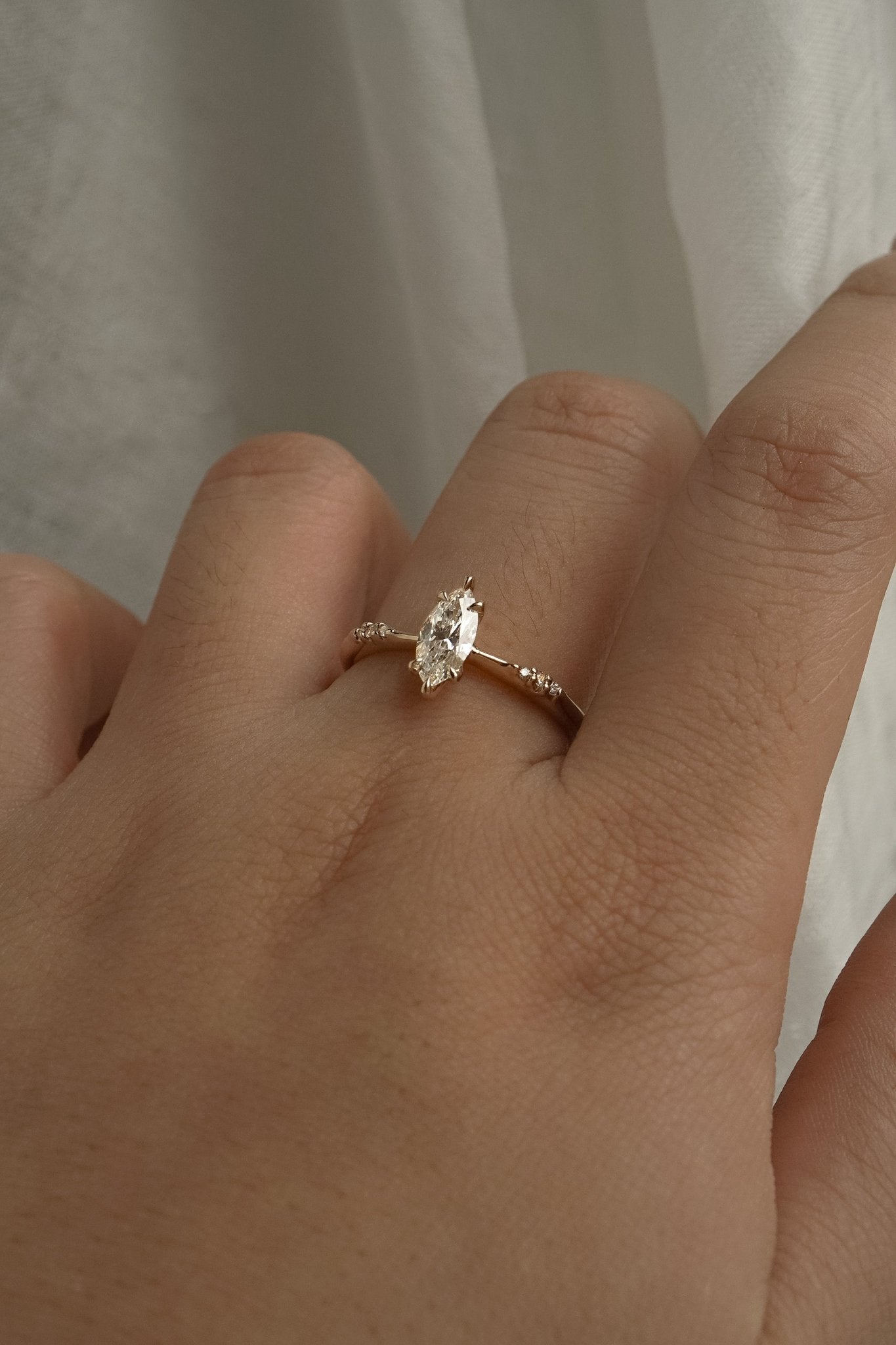 A delicate hand-carved one of a kind "Ilona" style engagement ring by Laurie Fleming Jewellery. The Ring features a 0.5ct marquise cut diamond, a tapered knife-edge band, and small diamond accents on the band shoulders.  The ring is worn on a hand posed in front of some draped gossamer silk fabric.