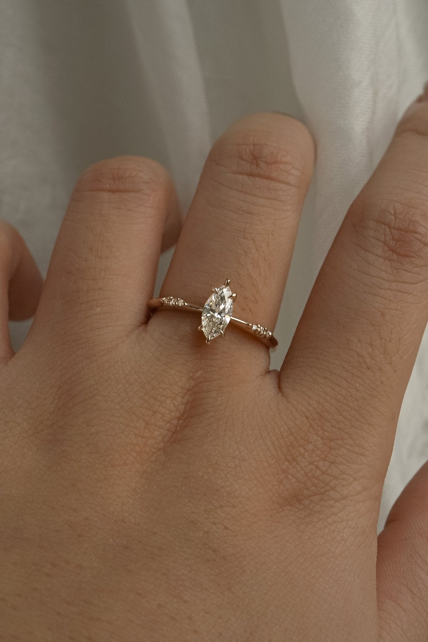 A delicate hand-carved one of a kind "Ilona" style engagement ring by Laurie Fleming Jewellery. The Ring features a 0.5ct marquise cut diamond, a tapered knife-edge band, and small diamond accents on the band shoulders. The ring is worn on a hand posed in front of some draped gossamer silk fabric.