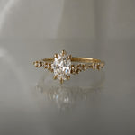 A one of a kind "Daphne" style engagement ring by Laurie Fleming jewellery, featuring a 0.70ct modified oval cut diamond with a unique glimmering sparkle, with diamond clusters on the band. The ring is situated on a medium grey background.