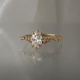 A one of a kind "Daphne" style engagement ring by Laurie Fleming jewellery, featuring a 0.70ct modified oval cut diamond with a unique glimmering sparkle, with diamond clusters on the band. The ring is situated on a medium grey background.