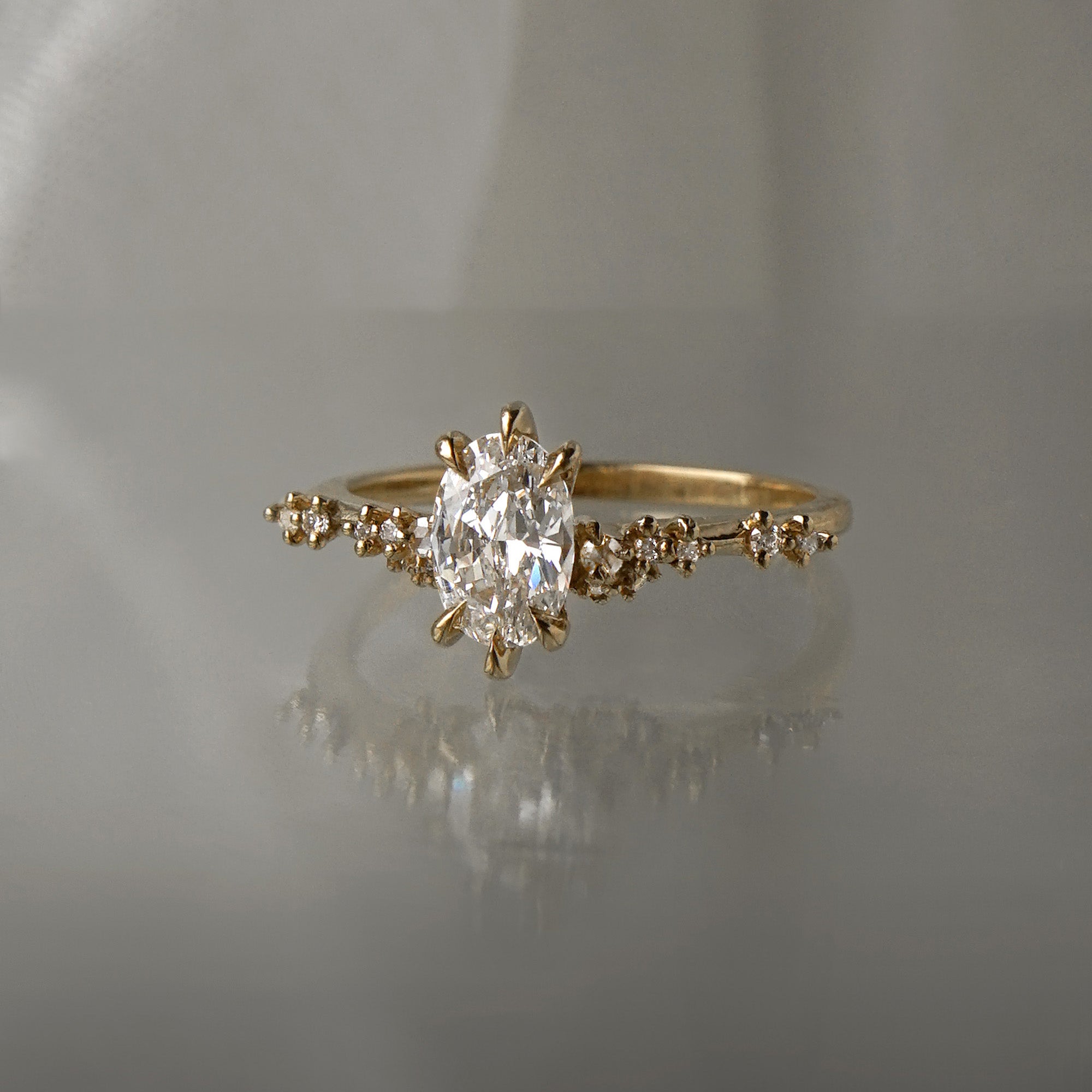 A one of a kind "Daphne" style engagement ring by Laurie Fleming jewellery, featuring a 0.70ct modified oval cut diamond with a unique glimmering sparkle, with diamond clusters on the band. The ring is situated on a medium grey background.