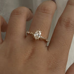 A one of a kind "Daphne" style engagement ring by Laurie Fleming jewellery, featuring a 0.70ct modified oval cut diamond with a unique glimmering sparkle, with diamond clusters on the band. The ring is worn on a hand that is posed in front of some draped gossamer silk fabric.