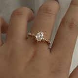 A one of a kind "Daphne" style engagement ring by Laurie Fleming jewellery, featuring a 0.70ct modified oval cut diamond with a unique glimmering sparkle, with diamond clusters on the band. The ring is worn on a hand that is posed in front of some draped gossamer silk fabric.