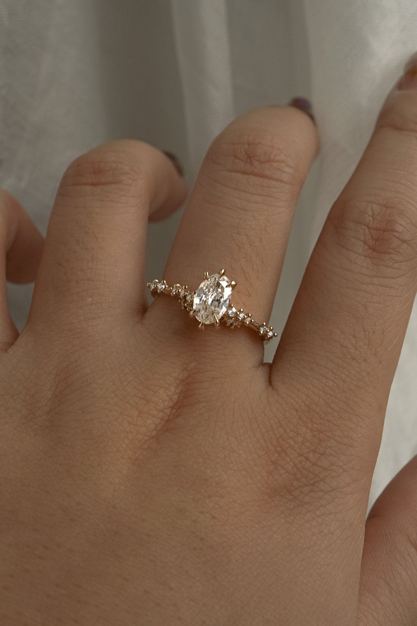 A one of a kind "Daphne" style engagement ring by Laurie Fleming jewellery, featuring a 0.70ct modified oval cut diamond with a unique glimmering sparkle, with diamond clusters on the band. The ring is worn on a hand that is posed in front of some draped gossamer silk fabric.