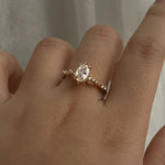A one of a kind "Daphne" style engagement ring by Laurie Fleming jewellery, featuring a 0.70ct modified oval cut diamond with a unique glimmering sparkle, with diamond clusters on the band. The ring is worn on a hand that is posed in front of some draped gossamer silk fabric.