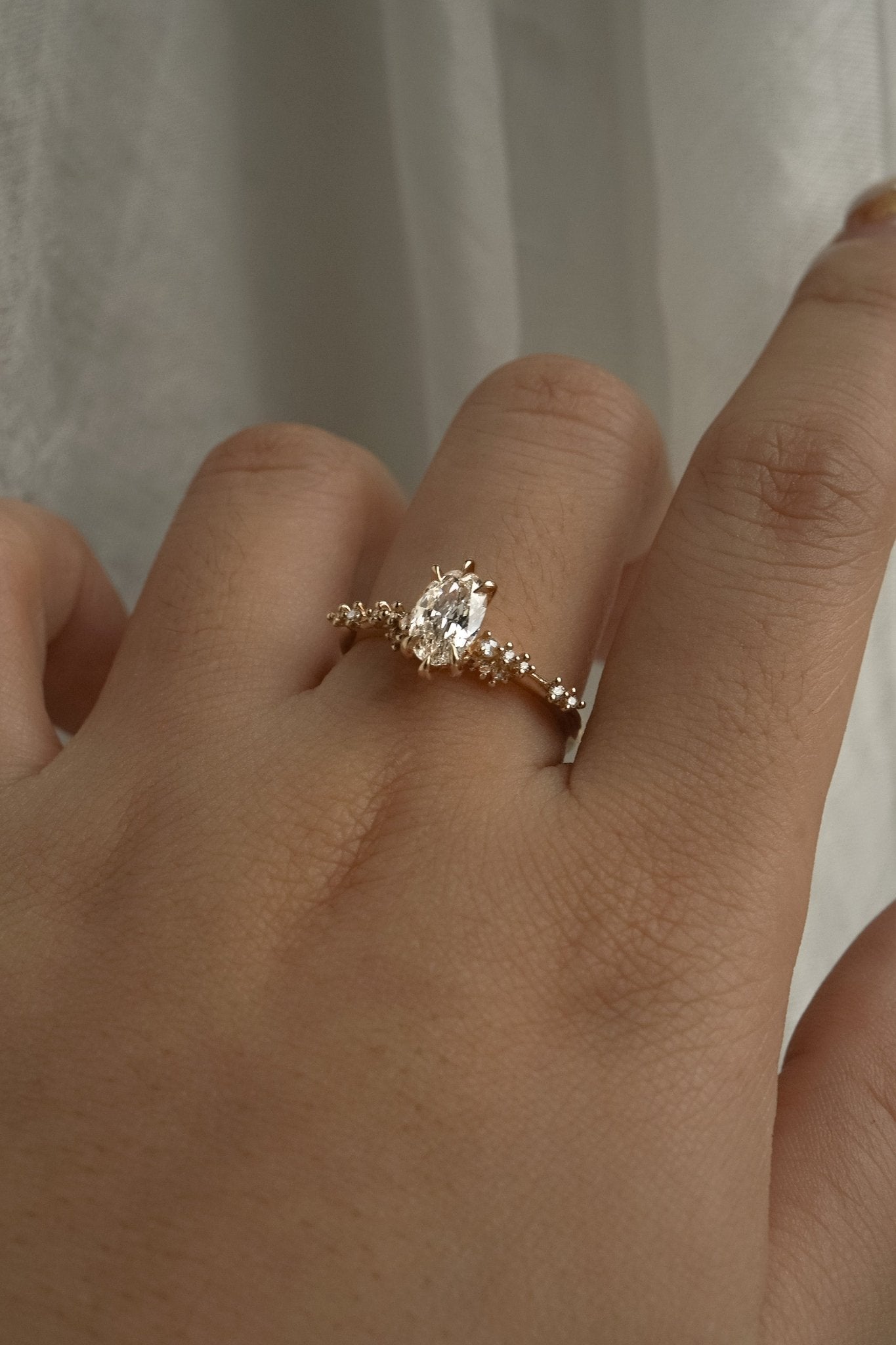 A one of a kind "Daphne" style engagement ring by Laurie Fleming jewellery, featuring a 0.70ct modified oval cut diamond with a unique glimmering sparkle, with diamond clusters on the band. The ring is worn on a hand that is posed in front of some draped gossamer silk fabric.