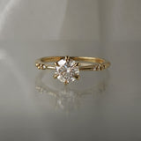 A delicate hand-carved one of a kind "Ilona" style engagement ring by Laurie Fleming Jewellery. The Ring features a 0.8ct round brilliant cut diamond, a tapered knife-edge band, and small diamond accents on the band shoulders. The ring is situated against a medium grey background.
