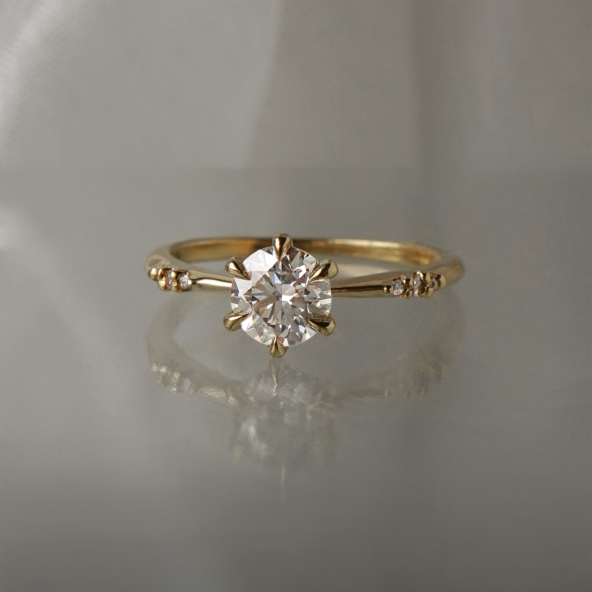 A delicate hand-carved one of a kind "Ilona" style engagement ring by Laurie Fleming Jewellery. The Ring features a 0.8ct round brilliant cut diamond, a tapered knife-edge band, and small diamond accents on the band shoulders. The ring is situated against a medium grey background.