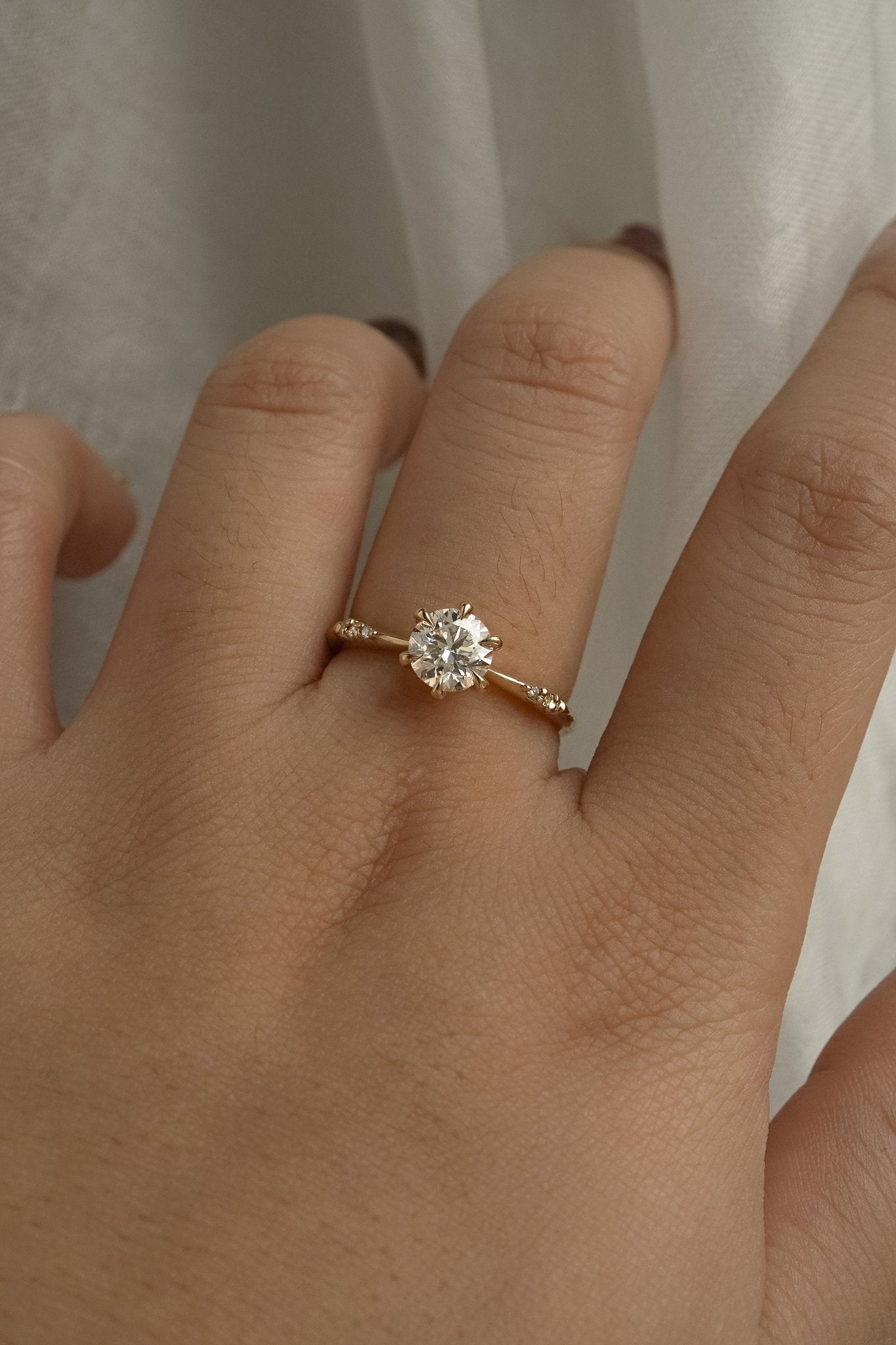 A delicate hand-carved one of a kind "Ilona" style engagement ring by Laurie Fleming Jewellery. The Ring features a 0.8ct round brilliant cut diamond, a tapered knife-edge band, and small diamond accents on the band shoulders. The ring is worn on a hand posed in front of some draped gossamer silk fabric.