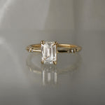 A one of a kind "Ilona" style engagement ring featuring a 0.92ct natural Emerald cut diamond held by four delicately hand-carved claw shaped prongs. The band tapers towards the centre and features diamonds on the shoulders. The ring is situated on a medium grey background.