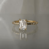 A one of a kind "Ilona" style engagement ring featuring a 0.92ct natural Emerald cut diamond held by four delicately hand-carved claw shaped prongs. The band tapers towards the centre and features diamonds on the shoulders. The ring is situated on a medium grey background.
