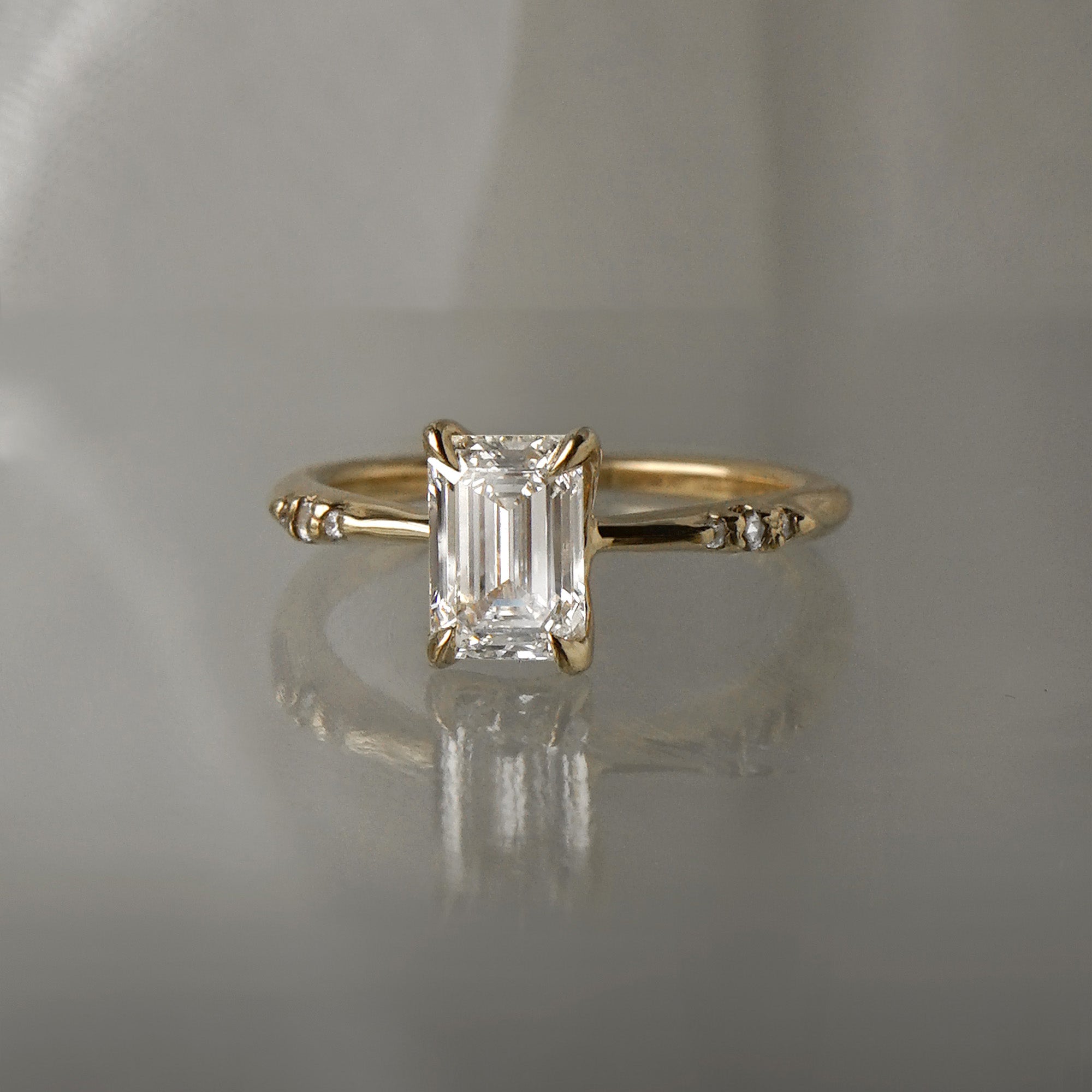 A one of a kind "Ilona" style engagement ring featuring a 0.92ct natural Emerald cut diamond held by four delicately hand-carved claw shaped prongs. The band tapers towards the centre and features diamonds on the shoulders. The ring is situated on a medium grey background.