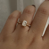 A one of a kind "Ilona" style engagement ring featuring a 0.92ct natural Emerald cut diamond held by four delicately hand-carved claw shaped prongs. The band tapers towards the centre and features diamonds on the shoulders. The ring is worn on a hand that is posed in front of some draped gossamer silk fabric.