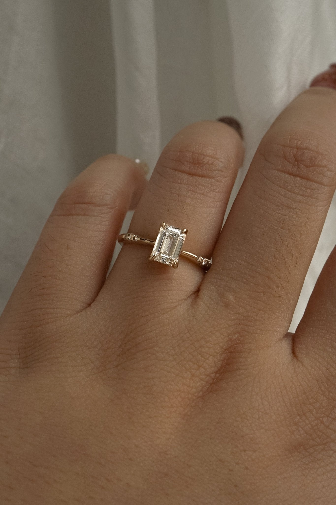 A one of a kind "Ilona" style engagement ring featuring a 0.92ct natural Emerald cut diamond held by four delicately hand-carved claw shaped prongs. The band tapers towards the centre and features diamonds on the shoulders. The ring is worn on a hand that is posed in front of some draped gossamer silk fabric.