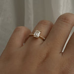 A one of a kind "Ilona" style engagement ring featuring a 0.92ct natural Emerald cut diamond held by four delicately hand-carved claw shaped prongs. The band tapers towards the centre and features diamonds on the shoulders. The ring is worn on a hand that is posed in front of some draped gossamer silk fabric.