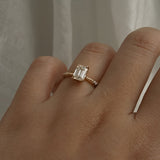 A one of a kind "Ilona" style engagement ring featuring a 0.92ct natural Emerald cut diamond held by four delicately hand-carved claw shaped prongs. The band tapers towards the centre and features diamonds on the shoulders. The ring is worn on a hand that is posed in front of some draped gossamer silk fabric.