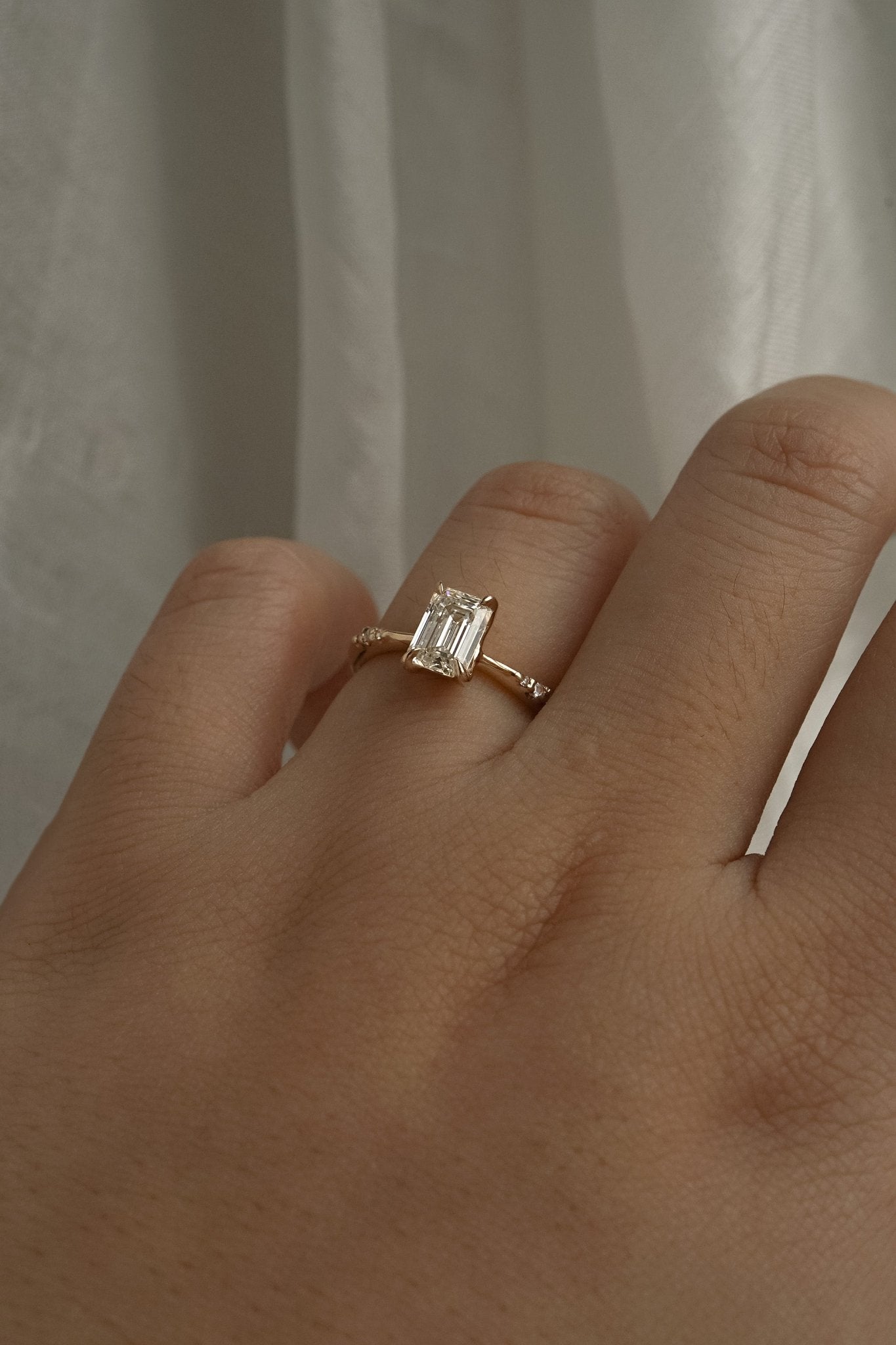 A one of a kind "Ilona" style engagement ring featuring a 0.92ct natural Emerald cut diamond held by four delicately hand-carved claw shaped prongs. The band tapers towards the centre and features diamonds on the shoulders. The ring is worn on a hand that is posed in front of some draped gossamer silk fabric.