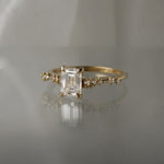 A delicate and ethereal hand-carved one of a kind "Daphne" style engagement ring by Laurie Fleming Jewellery. The ring features a 1.01ct emerald cut diamond with rose and brilliant cut diamond accents on the band in 14k yellow gold. The ring is situated against a medium grey background.