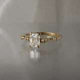 A delicate and ethereal hand-carved one of a kind "Daphne" style engagement ring by Laurie Fleming Jewellery. The ring features a 1.01ct emerald cut diamond with rose and brilliant cut diamond accents on the band in 14k yellow gold. The ring is situated against a medium grey background.
