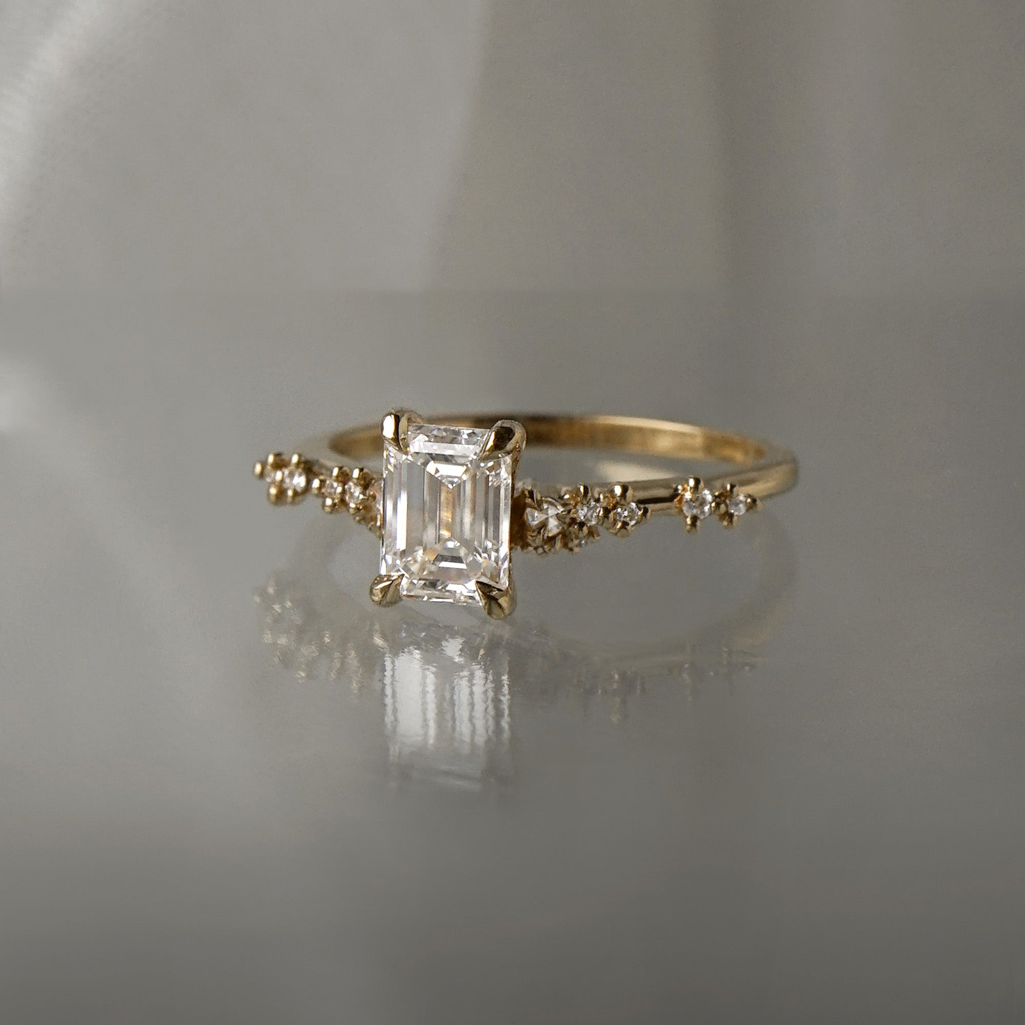 A delicate and ethereal hand-carved one of a kind "Daphne" style engagement ring by Laurie Fleming Jewellery. The ring features a 1.01ct emerald cut diamond with rose and brilliant cut diamond accents on the band in 14k yellow gold. The ring is situated against a medium grey background.