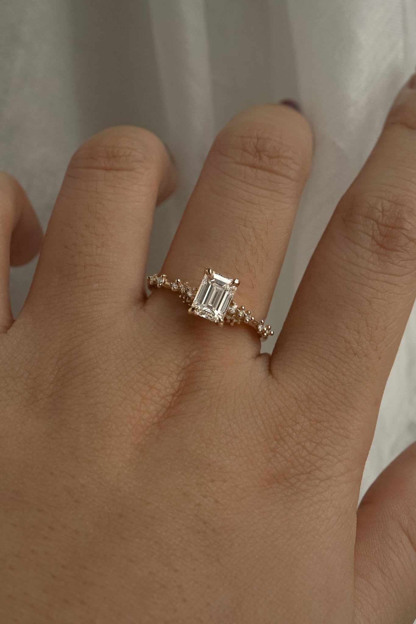 A delicate and ethereal hand-carved one of a kind "Daphne" style engagement ring by Laurie Fleming Jewellery. The ring features a 1.01ct emerald cut diamond with rose and brilliant cut diamond accents on the band in 14k yellow gold. The ring is worn on a hand posed in front of some draped gossamer silk fabric.