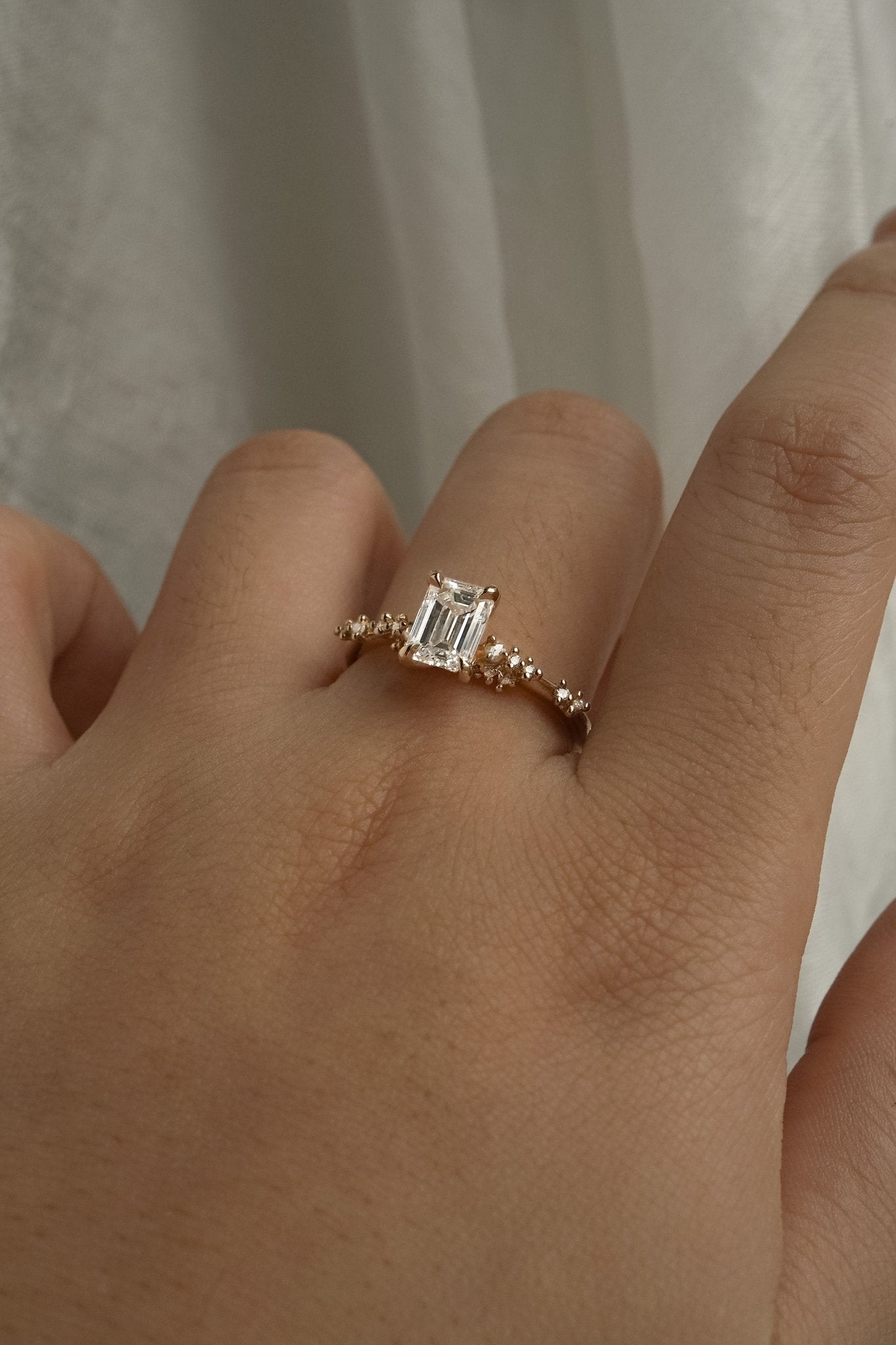 A delicate and ethereal hand-carved one of a kind "Daphne" style engagement ring by Laurie Fleming Jewellery. The ring features a 1.01ct emerald cut diamond with rose and brilliant cut diamond accents on the band in 14k yellow gold. The ring is worn on a hand posed in front of some draped gossamer silk fabric.
