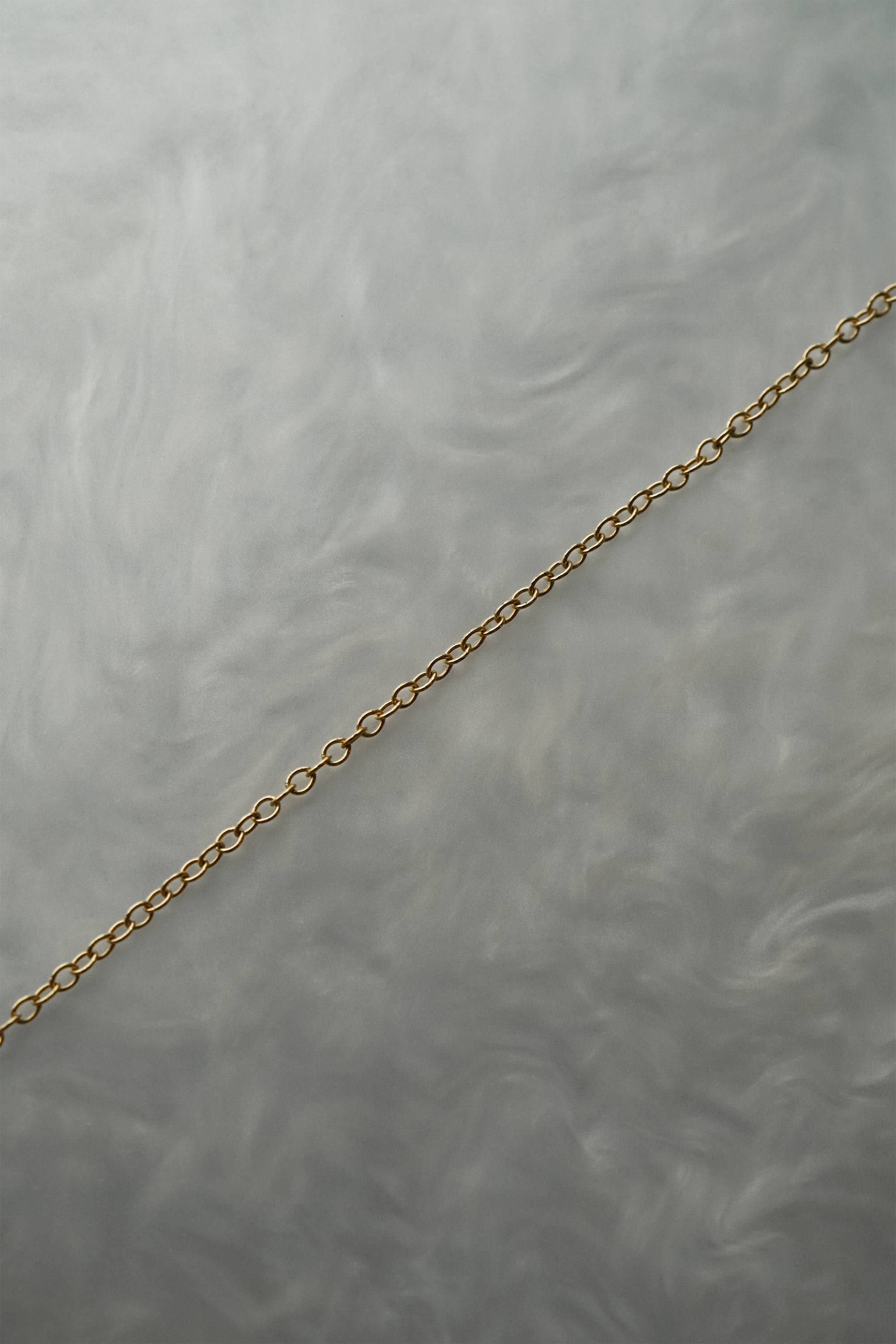 1.4mm cable chain with smooth oval links in solid 14k gold, perfect for wearing on its own or with charms or pendants. The chain is against a grey background with swirling patterns.