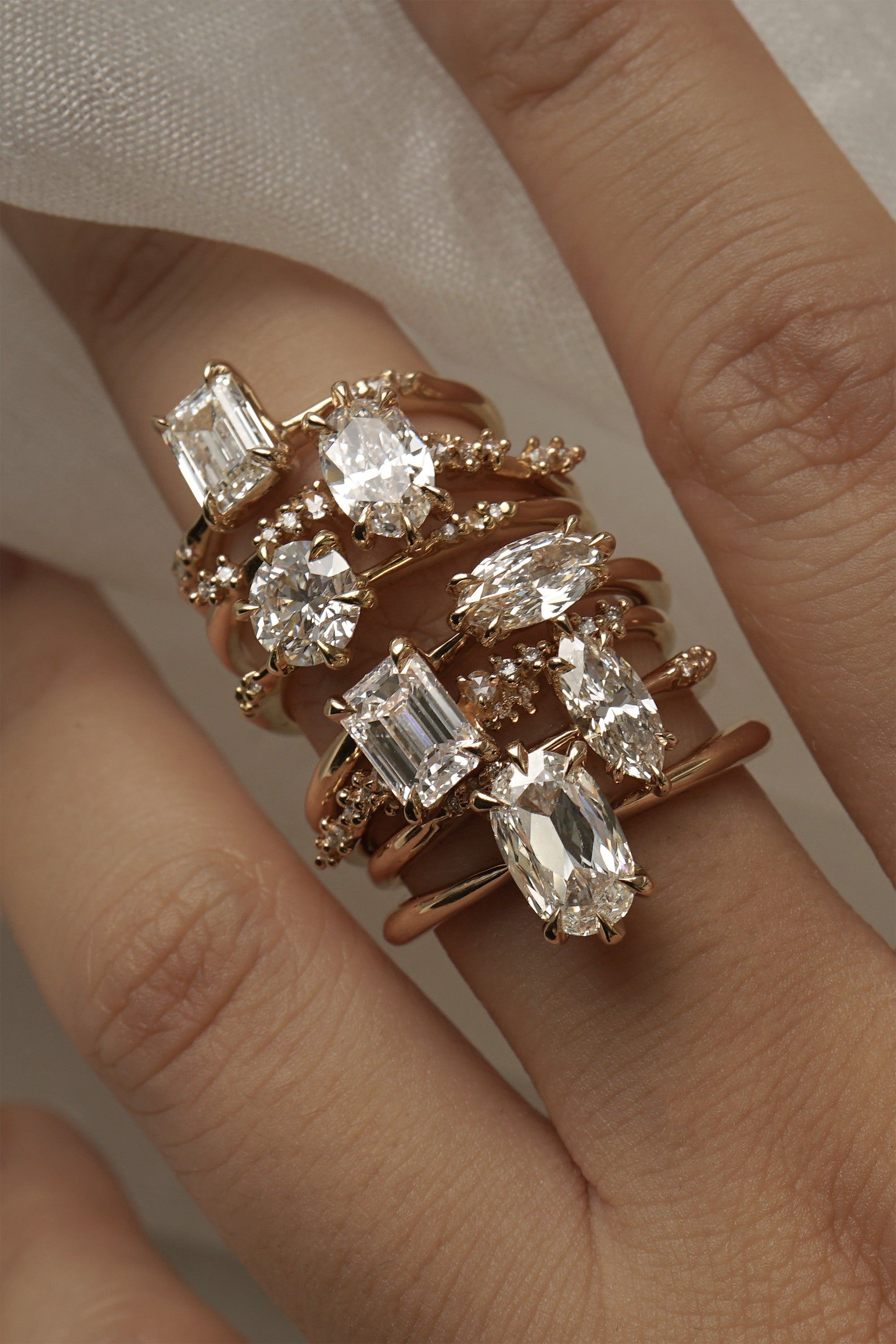 A hand with a stack of diamond engagement rings by Laurie Fleming Jewellery on the middle finger. From top to bottom: Ilona ring with emerald cut diamond, Daphne Ring with oval cut diamond, Ilona ring with round brilliant cut diamond, Aurelia ring with a moval (marquise/oval) diamond, Daphne ring with an emerald cut diamond, Ilona ring with a marquise cut diamond, Aurelia ring with an oval cut diamond.