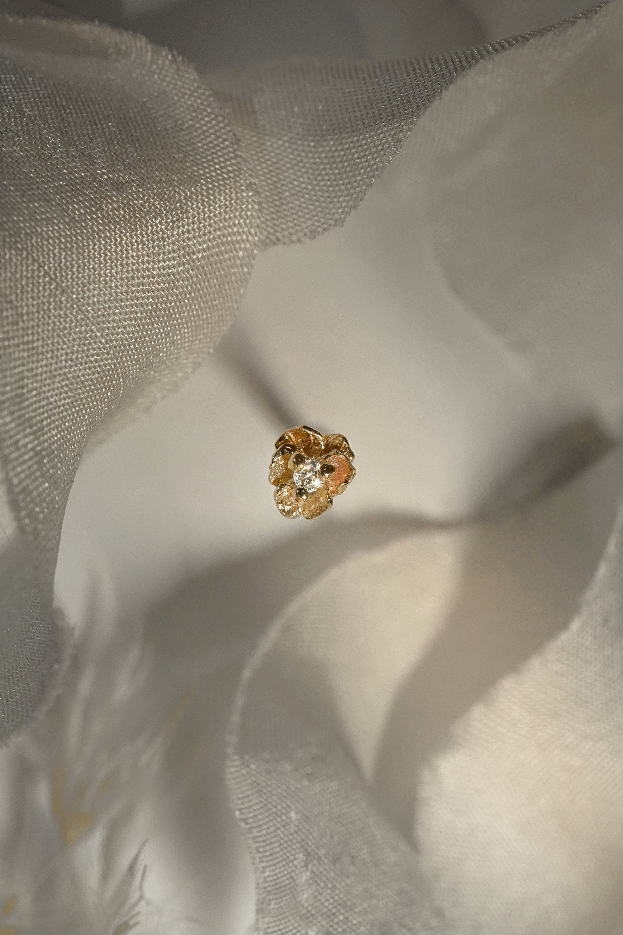 A product photo of a Pansy stud by Laurie Fleming Jewellery. It features a hand-carved miniature Pansy flower in solid 14k gold with a round brilliant cut diamond centre. The diamond can be swapped out for any birthstone. The background is a light grey with bits of raw silk at the top and bottom of the frame.