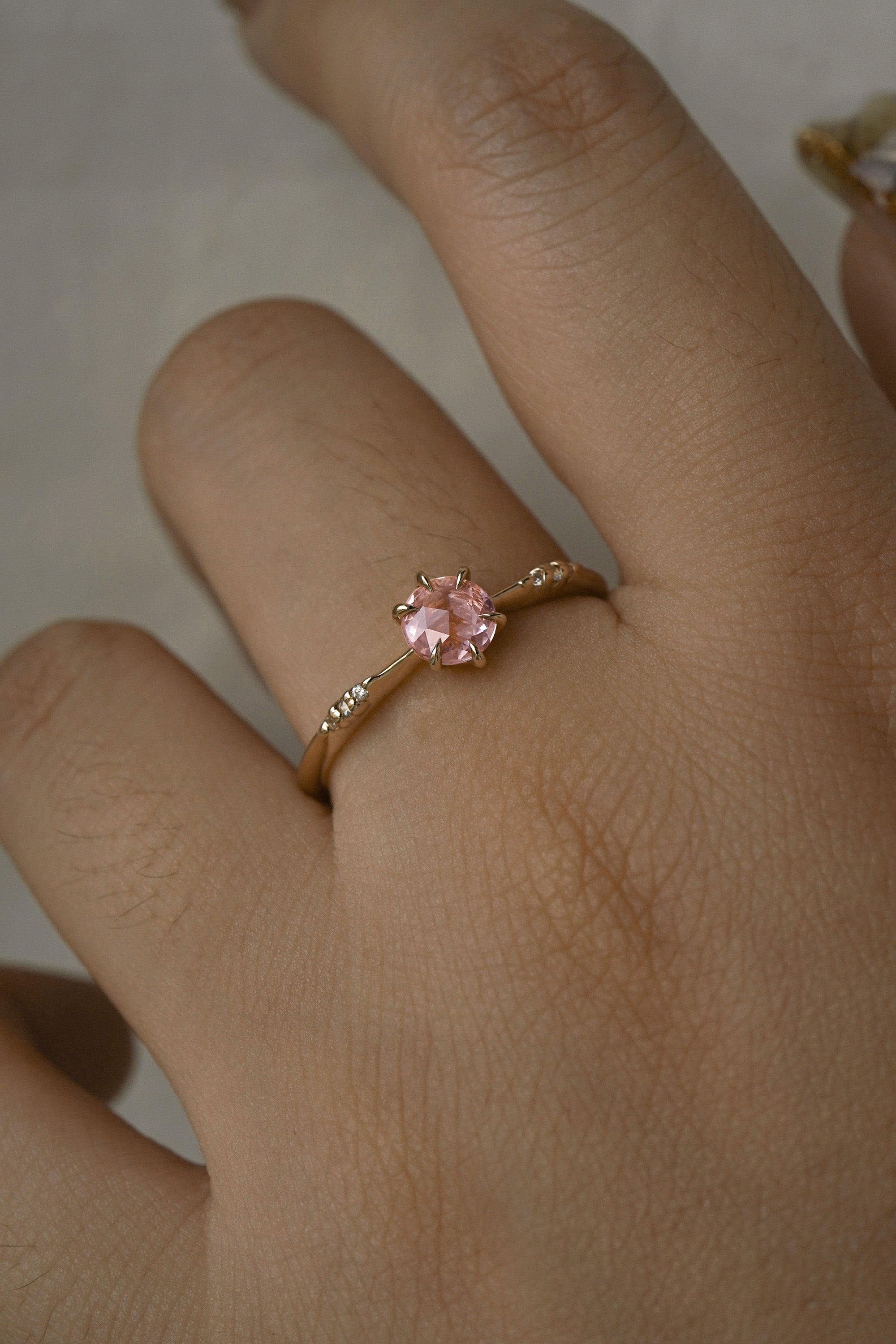 A delicate hand-carved one of a kind "Ilona" style engagement ring by Laurie Fleming Jewellery. The ring features a rose cut pink sapphire centre, a tapered knife-edge band, and small diamond accents on the band shoulders. The ring is worn on a hand against a light grey background.