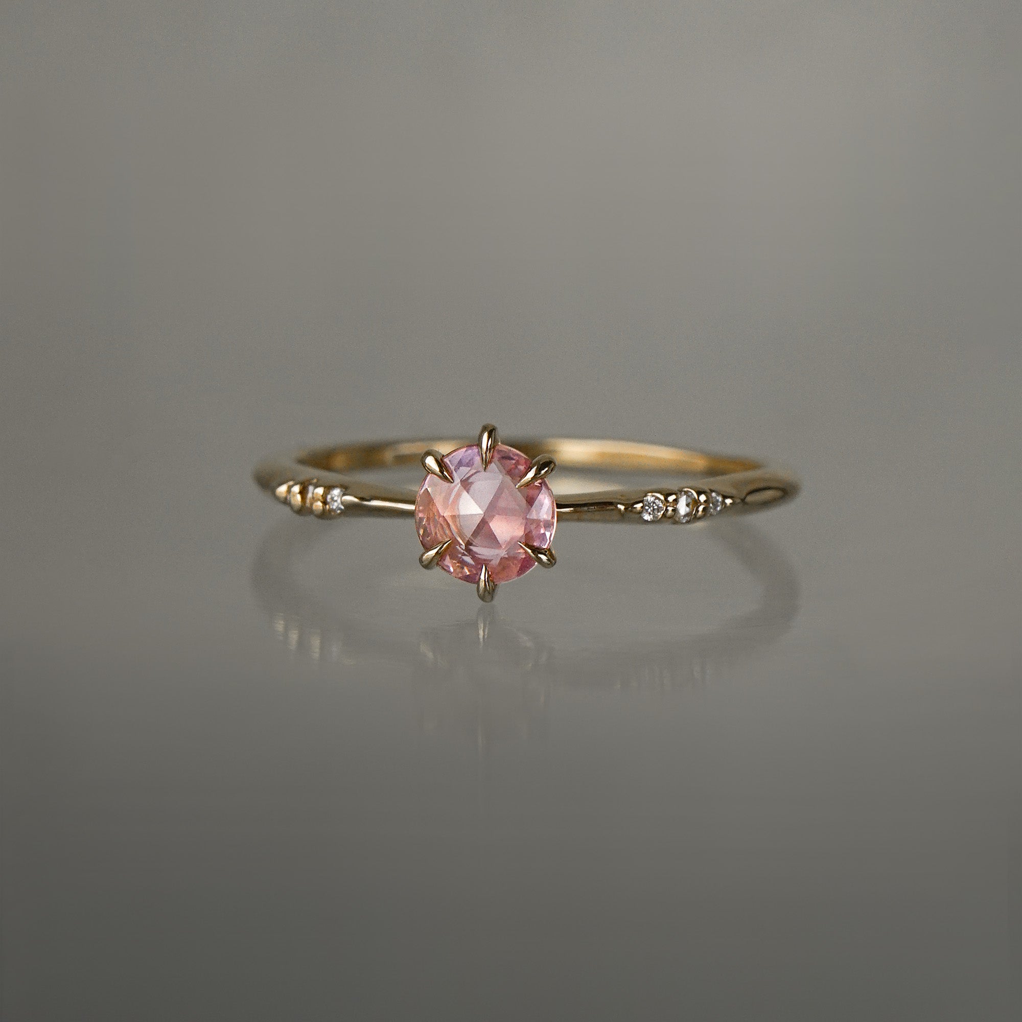 A delicate hand-carved one of a kind "Ilona" style engagement ring by Laurie Fleming Jewellery. The ring features a rose cut pink sapphire centre, a tapered knife-edge band, and small diamond accents on the band shoulders.  The ring is situated against a medium grey background.
