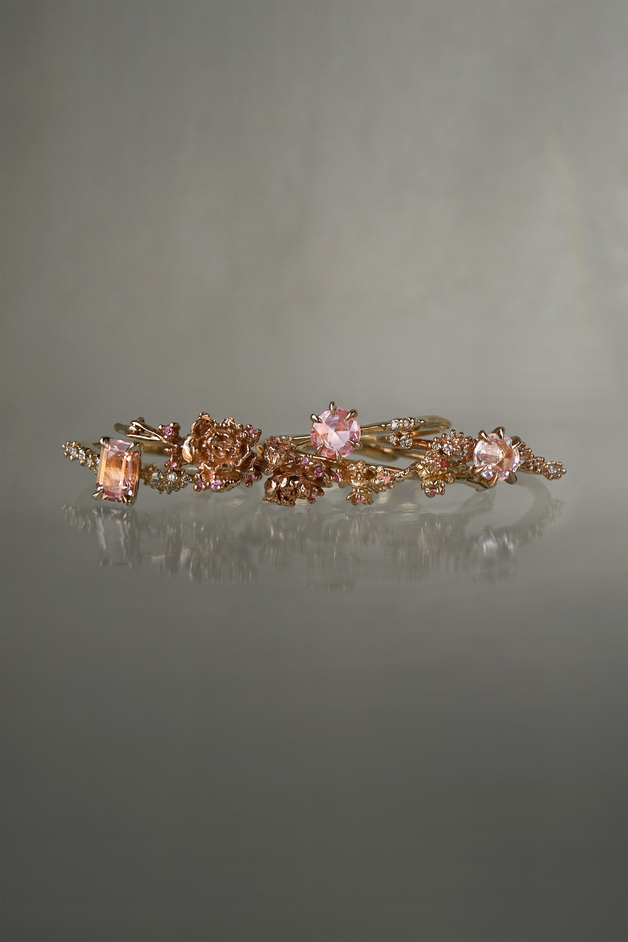 A photo of three rings and two hoops made by Laurie Fleming Jewellery against a medium grey background. From left to right: A "Daphne" style engagement ring featuring an emerald cut peach sapphire, two one of a kind "Asrai Garden" hoops featuring hand-carved miniature flowers, an "Ilona" style engagement ring with a round rose cut pink sapphire, and a "Daphne" style engagement ring with a round rose cut pale pink sapphire.