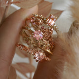 A photo of three rings and two hoops made by Laurie Fleming Jewellery held between index finger and thumb, including:  A "Daphne" style engagement ring featuring an emerald cut peach sapphire, two one of a kind "Asrai Garden" hoops featuring hand-carved miniature flowers, an "Ilona" style engagement ring with a round rose cut pink sapphire, and a "Daphne" style engagement ring with a round rose cut pale pink sapphire. There are leaves and feathers in shades of soft beige and pink surrounding the hand,