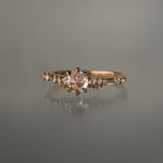 A delicate and ethereal hand-carved one of a kind "Daphne" style engagement ring by Laurie Fleming Jewellery. The ring features a rose cut pale pink sapphire centre, with clusters of rose and brilliant cut diamond accents on the band in solid 14k rose gold. The ring is situated against a medium grey background.