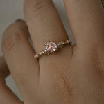 A delicate and ethereal hand-carved one of a kind "Daphne" style engagement ring by Laurie Fleming Jewellery. The ring features a rose cut pale pink sapphire centre, with clusters of rose and brilliant cut diamond accents on the band in solid 14k rose gold. The ring is worn on a hand against a light grey background.