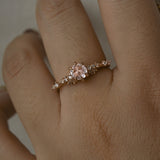 A delicate and ethereal hand-carved one of a kind "Daphne" style engagement ring by Laurie Fleming Jewellery. The ring features a rose cut pale pink sapphire centre, with clusters of rose and brilliant cut diamond accents on the band in solid 14k rose gold. The ring is worn on a hand against a light grey background.