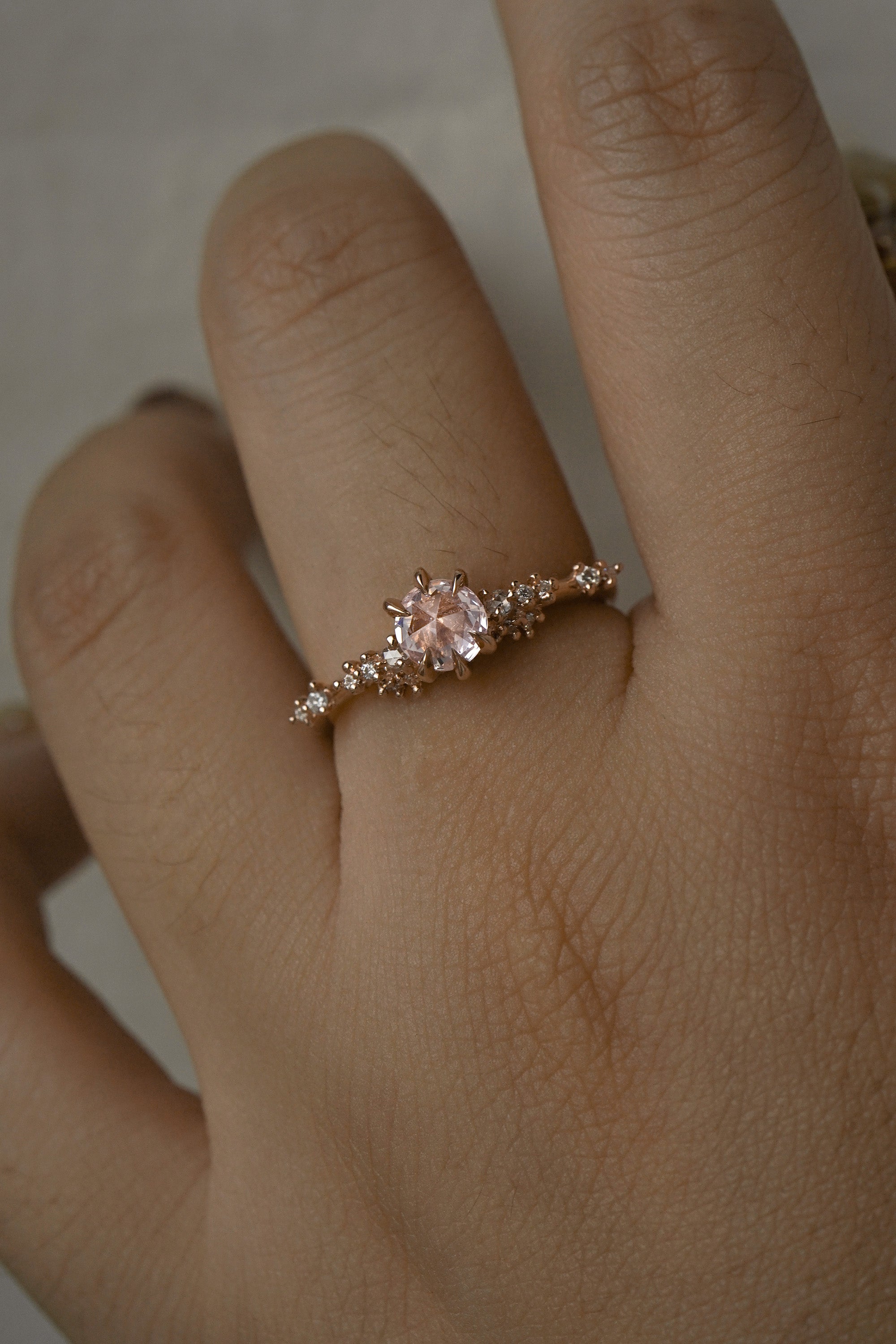 A delicate and ethereal hand-carved one of a kind "Daphne" style engagement ring by Laurie Fleming Jewellery. The ring features a rose cut pale pink sapphire centre, with clusters of rose and brilliant cut diamond accents on the band in solid 14k rose gold. The ring is worn on a hand against a light grey background.
