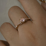 A delicate and ethereal hand-carved one of a kind "Daphne" style engagement ring by Laurie Fleming Jewellery. The ring features a rose cut pale pink sapphire centre, with clusters of rose and brilliant cut diamond accents on the band in solid 14k rose gold. The ring is worn on a hand against a light grey background.
