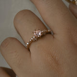 A delicate and ethereal hand-carved one of a kind "Daphne" style engagement ring by Laurie Fleming Jewellery. The ring features a rose cut pale pink sapphire centre, with clusters of rose and brilliant cut diamond accents on the band in solid 14k rose gold. The ring is worn on a hand against a light grey background.