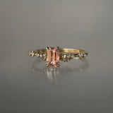 A delicate and ethereal hand-carved one of a kind "Daphne" style engagement ring by Laurie Fleming Jewellery. The ring features an emerald cut peach-pink Montana sapphire centre, with clusters of rose and brilliant cut diamond accents on the band in solid 14k yellow gold. The ring is situated against a medium grey background.