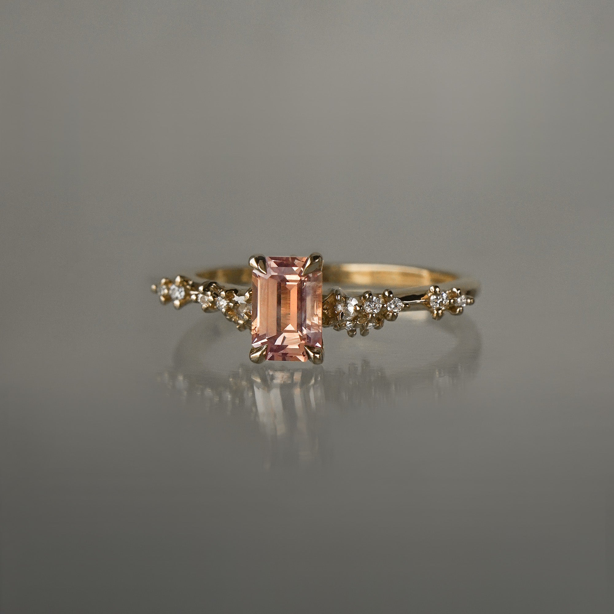 A delicate and ethereal hand-carved one of a kind "Daphne" style engagement ring by Laurie Fleming Jewellery. The ring features an emerald cut peach-pink Montana sapphire centre, with clusters of rose and brilliant cut diamond accents on the band in solid 14k yellow gold. The ring is situated against a medium grey background.