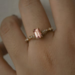 A delicate and ethereal hand-carved one of a kind "Daphne" style engagement ring by Laurie Fleming Jewellery. The ring features an emerald cut peach-pink Montana sapphire centre, with clusters of rose and brilliant cut diamond accents on the band in solid 14k yellow gold. The ring is worn on a hand against a light grey background.