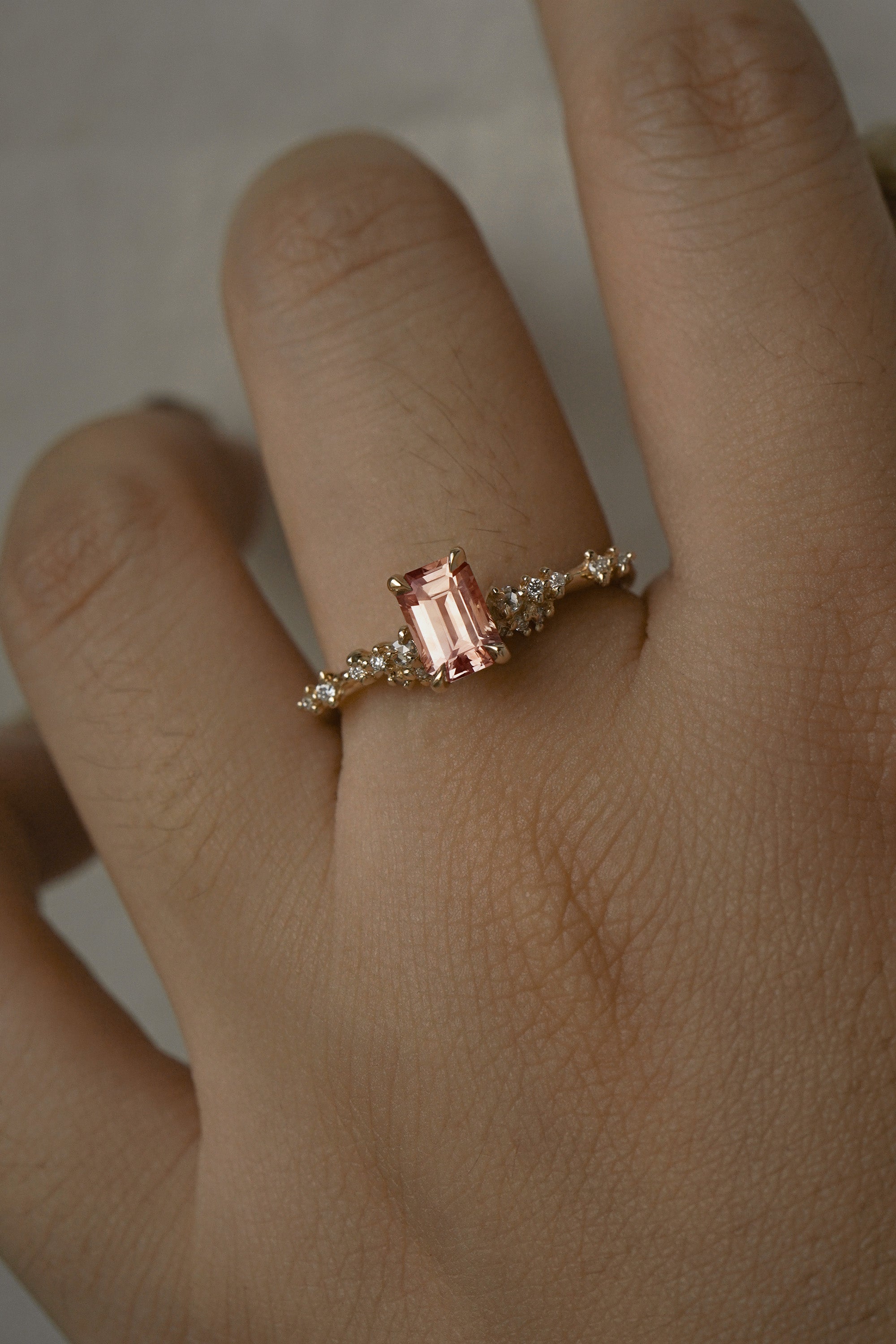 A delicate and ethereal hand-carved one of a kind "Daphne" style engagement ring by Laurie Fleming Jewellery. The ring features an emerald cut peach-pink Montana sapphire centre, with clusters of rose and brilliant cut diamond accents on the band in solid 14k yellow gold. The ring is worn on a hand against a light grey background.