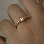 A delicate and ethereal hand-carved one of a kind "Daphne" style engagement ring by Laurie Fleming Jewellery. The ring features an emerald cut peach-pink Montana sapphire centre, with clusters of rose and brilliant cut diamond accents on the band in solid 14k yellow gold. The ring is worn on a hand against a light grey background.