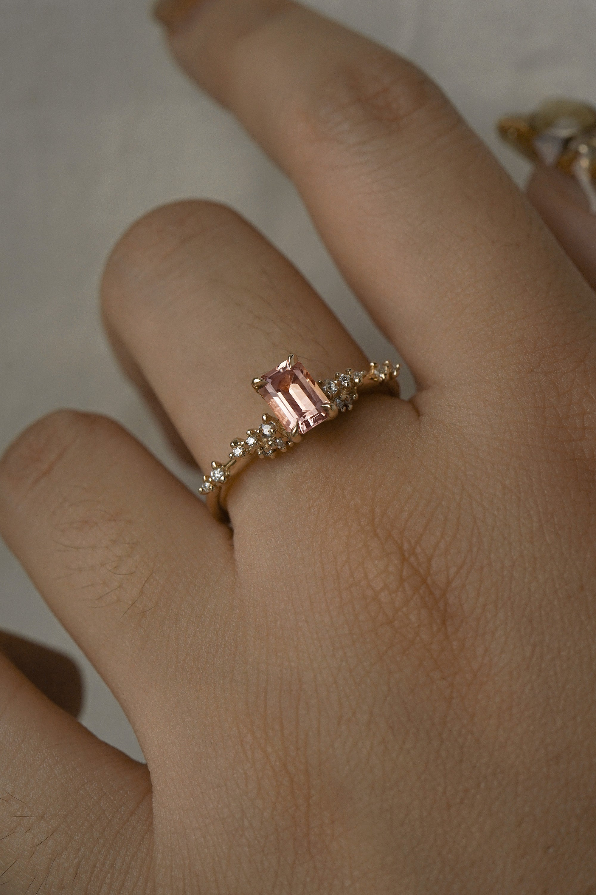 A delicate and ethereal hand-carved one of a kind "Daphne" style engagement ring by Laurie Fleming Jewellery. The ring features an emerald cut peach-pink Montana sapphire centre, with clusters of rose and brilliant cut diamond accents on the band in solid 14k yellow gold. The ring is worn on a hand against a light grey background.