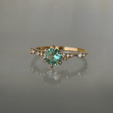 A delicate and ethereal hand-carved one of a kind "Daphne" style engagement ring by Laurie Fleming Jewellery. The ring features a round brilliant cut light green sapphire centre, with clusters of rose and brilliant cut diamond accents on the band in 14k yellow gold. The ring is situated against a medium grey background.