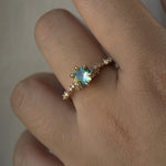 A delicate and ethereal hand-carved one of a kind "Daphne" style engagement ring by Laurie Fleming Jewellery. The ring features a round brilliant cut light green sapphire centre, with clusters of rose and brilliant cut diamond accents on the band in 14k yellow gold. The ring is worn on a hand against a light grey background.