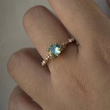 A delicate and ethereal hand-carved one of a kind "Daphne" style engagement ring by Laurie Fleming Jewellery. The ring features a round brilliant cut light green sapphire centre, with clusters of rose and brilliant cut diamond accents on the band in 14k yellow gold. The ring is worn on a hand against a light grey background.